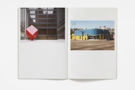 An open book showing two photographic images. The left page features a large red cube sculpture in front of a modern building. The right page displays a yellow building with a blue and white checkered pattern on its upper facade, situated on a street corner. 