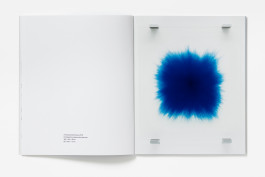 A spread from Idris Khan: Words Beneath Words with a blank left page showing artwork details, and a right page featuring a vibrant blue rubber stamp painting layered on glass, with wall fixings in the corners.